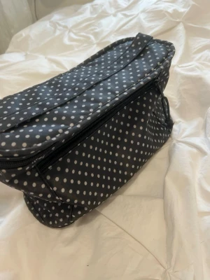 Svart necessär med vita prickar - Säljer en svart necessär med vita prickar och dragkedja. Väskan har ett praktiskt handtag på locket och en liten spegel på insidan. Perfekt för smink eller småprylar. Tillverkad i ett tåligt tygmaterial.