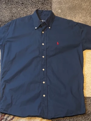 Mörkblå skjorta från Ralph Lauren - Klassisk mörkblå skjorta från Ralph Lauren med kort ärm och knappar framtill. Skjortan har en liten röd broderad logga på bröstet och button-down krage. Perfekt för en stilren och avslappnad look.