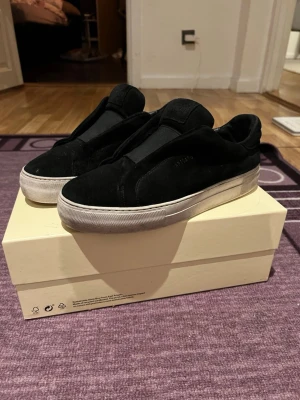Svarta sneakers från Axel Arigato - Säljer ett par svarta sneakers från Axel Arigato i mocka med vit platt sula. Skorna har minimalistisk design utan snörning och diskret logga på sidan. Perfekta för dig som gillar stilrena och moderna skor. Strl 41