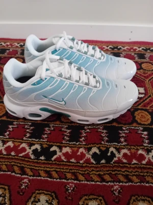 Nike Air Max Plus vita/blå sneakers - Nike Air Max Plus sneakers i vitt och ljusblått med ikoniska vågformade linjer och synlig Air Max-dämpning. Ovandel i mesh och syntet, vita skosnören och detaljer i turkos. Snygg och sportig siluett med svart yttersula och TN-logga under sulan.
