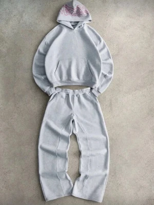 Grått loungewear-set med hoodie - Mysigt loungewear-set i ljusgrått med oversized hoodie och matchande byxor. Hoodien har magficka och huva med rosa grafiskt tryck. Byxorna har resår i midjan och raka ben. Perfekt för chill dagar hemma.