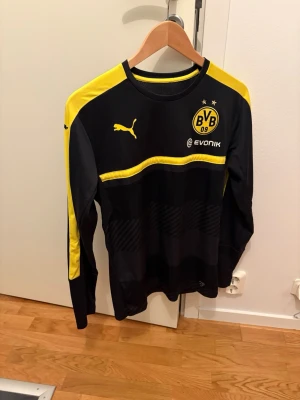 BVB långärmad träningströja Puma - Snygg svart och gul långärmad träningströja från Puma med Borussia Dortmunds BVB-logga och Evonik-tryck på bröstet. Tröjan har gula detaljer på axlar och ärmar samt rund hals. Perfekt för fotbollsträning eller som supporterplagg.