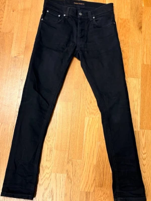 Nudie Jeans - Säljer ett par svarta jeans från Nudie Jeans med klassisk femficksdesign och smal passform. Jeansen har svarta sömmar och subtila detaljer på bakfickorna. Perfekta för en stilren och modern look. W30 L32