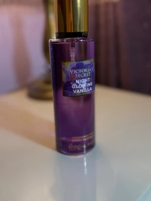 Victorias Secret Night Glowing Vanilla - En parfymmist från Victorias Secret i doften Night Glowing Vanilla. Flaskan är lila med guldigt lock och har en snygg etikett framtill. Innehåller 250 ml och har en elegant, cylinderformad design som känns lyxig och modern.