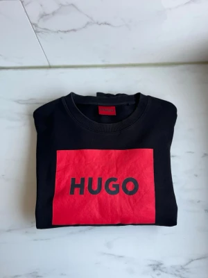 Svart sweatshirt från Hugo med röd logga - Cool svart sweatshirt från Hugo med stor röd ruta och tydlig logga på bröstet. Tröjan har rund halsringning och långa ärmar. Storlek L fits L/ stor M, fint skick ! 