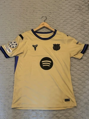 Gul FC Barcelona matchtröja - Snygg gul FC Barcelona matchtröja med svarta detaljer och tryck, inklusive klubbmärke, Spotify-logga och Champions League-märke på ärmen. Tröjan har blå och svarta ränder vid krage och ärmslut. Tillverkad i lätt och ventilerande material, perfekt för fotbollsträning eller match.
