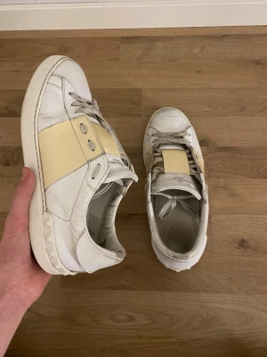 Valentino vita sneakers med beige detalj - Snygga vita sneakers från Valentino med en bred beige panel över mitten och silverfärgade nitar på sidorna. Skorna är låga och har klassisk snörning. Tillverkade i läder och levereras med originalkartong och dustbag.