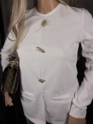 Vit kavaj med guldknappar - Stilren vit kavaj med unika guldknappar och dekorativ brosch upptill. Kavajen har rak passform, rund halsringning och två fickor framtill. Materialet är lätt ribbat och ger en modern känsla. Perfekt för dig som vill sticka ut med detaljer.