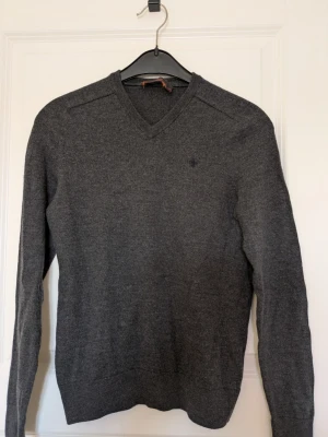 Mörkgrå stickad tröja, Morris. med V-ringning - Mörkgrå stickad tröja med V-ringad hals och diskret broderad logga på bröstet. Tröjan har långa ärmar och ribbade muddar vid ärmslut och nederkant. Perfekt för lager på lager och enkel att matcha till olika outfits. Mått: höjd 58 cm, axelbredd 48 cm.