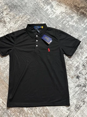 Svart Polo Ralph Lauren pikétröja - Klassisk svart pikétröja från Polo Ralph Lauren med röd broderad logga på bröstet och knappar framtill. Tröjan har korta ärmar och mjukt bomullsmaterial som känns skönt mot huden. Perfekt för en stilren och avslappnad look.