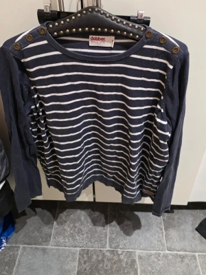 Marinblå randig tröja från Dobber - Snygg marinblå tröja från Dobber med vita horisontella ränder. Tröjan har rund halsringning och dekorativa knappar på axlarna. Långärmad och i mjukt material, perfekt för en avslappnad stil.