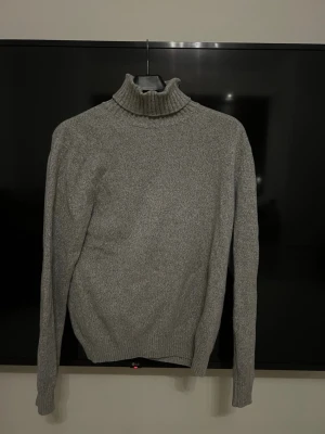 Grå polotröja i stickat material - Säljer en stilren grå stickad tröja med hög krage och långa ärmar. Tröjan har ribbade muddar vid ärmslut och nederkant, vilket ger en snygg passform. Perfekt för dig som gillar en enkel men trendig look.