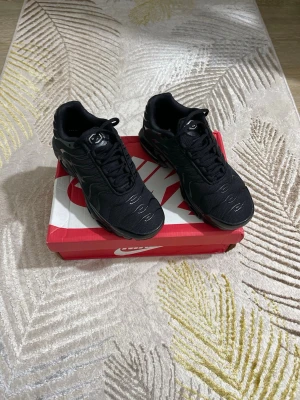 Nike Air Max Plus helsvarta sneakers - Snygga helsvarta Nike Air Max Plus sneakers med mesh- och syntetöverdel, ikoniska vågformade detaljer och synlig Air Max-dämpning i sulan. Perfekta för dig som gillar sportig och stilren look. Klassisk Nike-logga på tungan och sidan.