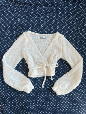 Vit omlottstickad topp Hollister - Säljer en vit stickad omlottopp från Hollister med långa ärmar och knytband i midjan. Toppen är croppad och har en djup v-ringning. Materialet är mjukt och fluffigt, perfekt för lager på lager och en chill look. Måtten finns. 