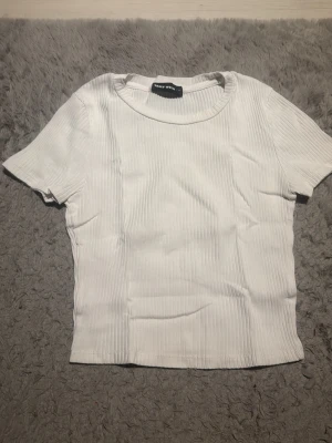 Vit ribbad t-shirt från Trashy White - Snygg vit ribbad t-shirt från Trashy White med korta ärmar och rund halsringning. T-shirten har en figurnära passform och är tillverkad i ett stretchigt material som ger en bekväm känsla. Perfekt att matcha med jeans eller kjol.