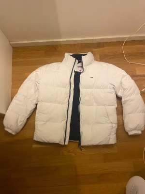 Vit pufferjacka från Tommy Jeans - Säljer en snygg vit pufferjacka från Tommy Jeans med mörkblå foder och diskret logga på bröstet. Jackan har hög krage, dragkedja framtill och elastiska muddar vid ärmslut. Perfekt för kalla dagar och har en clean, modern look.