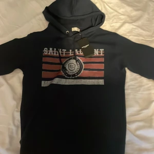 Saint Laurent hoodie - Snygg mörkblå hoodie från Saint Laurent med tryck i rött och vitt på bröstet. Tröjan har huva och snörning. Storlek S, Endast testad, modell 175, 60 kg, fraktar samma dag, skick 10/10, skriv vid funderingar eller intresse av köp!🙌🏻