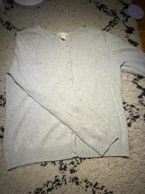Ljusgrå kofta med knappar från H&M - Ljusgrå, mjuk och stilren kofta från H&M med rund hals och långa ärmar. Koftan har ribbade muddar och knäppning framtill med matchande grå knappar. Perfekt för lager-på-lager och chill dagar.