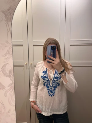 Vit blus med blå broderi från Indiska - Supersöt vit blus från Indiska i storlek S med blått broderat mönster framtill. Blusen har långa ärmar, v-ringning med knyt och tofsdetalj. Materialet är lätt och luftigt, perfekt för vår och sommar. Passformen är normal och den är köpt på Sellpy.