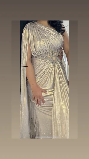 Champagnefärgad långklänning med slits - Säljer en elegant champagnefärgad långklänning med glittrigt tyg och draperad design. Klänningen har oneshoulder, vackra pärldetaljer i midjan och en slits på sidan. Materialet är skimrande och ger en lyxig känsla, perfekt för bal eller fest. Flödig cape på ena axeln för extra drama.