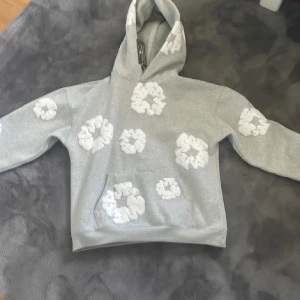 Grå hoodie från Denim Tears med blommor - Grå hoodie från Denim Tears med vita blommiga tryck över hela tröjan. Klassisk huva och känguruficka framtill. Mjuk och skön bomullskvalitet, perfekt för en chill och avslappnad stil.