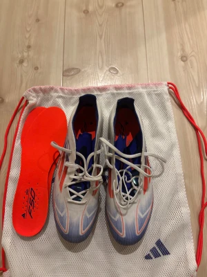 Adidas F50 elit fotbollsskor i vitt och blått - Säljer ett par Adidas F50 elit fotbollsskor i vitt med blå och röda detaljer. Skorna har snörning och en platt sula, samt en sportig design med spetsig tå. Materialet är syntet och de kommer med en extra röd innersula och en vit mesh-påse med Adidas-logga. Aldrig använda