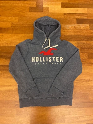 Mörkgrå Hollister hoodie M - Snygg och skön mörkgrå hoodie från Hollister med stor vit och röd logga på bröstet. Tröjan har huva med vit snörning och en klassisk känguruficka framtill. Mjuk insida i fleece och ribbade muddar vid ärmslut och nederkant. Perfekt för chill dagar.