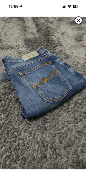 Nudie Jeans blå straight fit jeans - Säljer ett par klassiska blå jeans från Nudie Jeans med snygga kontrastsömmar och ikonisk broderad bakficka. Jeansen har rak passform och normal midja, tillverkade i slitstarkt bomullsdenim. Perfekta för dig som gillar en tidlös och avslappnad stil.