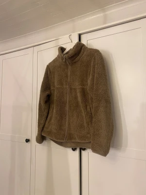 Beige teddyjacka från 157, storlek S - Mysig teddyjacka i beige från 157, perfekt för kalla dagar. Jackan har hög krage, hel dragkedja framtill och är tillverkad i fluffigt syntetmaterial. Passformen är normal och jackan har långa ärmar. Enkel och stilren design som är lätt att matcha.