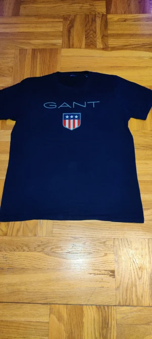 Marinblå GANT t-shirt med tryck - Snygg marinblå t-shirt från GANT med stort tryck framtill och klassiskt GANT-emblem med stjärnor och ränder. T-shirten har rund hals, korta ärmar och är gjord i mjuk bomull. Perfekt för en clean och sportig look.