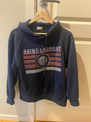 Mörkblå Saint Laurent hoodie - Säljer en mörkblå hoodie med tryck i vitt och rött framtill. Den är använd endast ett fåtal gånger. Storlek M men passar som XS/S, skulle säga 164-174. Tröjan har huva med snörning, stor magficka och långa ärmar. Materialet är mjuk bomull och passformen är normal. OBS lnte origlnal. Hör av dig vid frågor elr liknande🙌