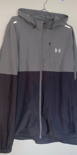 Grå och svart vindjacka Under Armour XL - Snygg vindjacka från Under Armour i storlek XL. Jackan är tvåfärgad med grått upptill och svart nedtill, har huva med snörning och hel dragkedja framtill. Logga på bröstet och praktisk bröstficka med dragkedja. Perfekt för aktiva dagar.