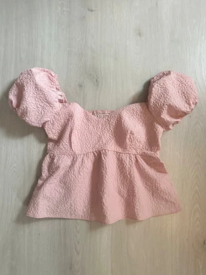 Rosa blus med puffärmar - Supersöt ljusrosa blus med puffiga korta ärmar och strukturerat tyg. Blusen har en v-ringad hals och smock i ryggen för extra komfort. Den har en volang nedtill som ger en markerad midja. Blusen är kortare i modellen och i topp skick, aldrig använd! 