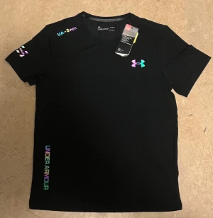 Svart Under Armour t- shirt - Snygg svart t-shirt från Under Armour med korta ärmar och coola färgglada loggor på bröstet, axeln och nederkanten. Tillverkad i mjuk polyester som andas, perfekt för dig som gillar sportig stil och vill sticka ut med diskreta men färgstarka detaljer.