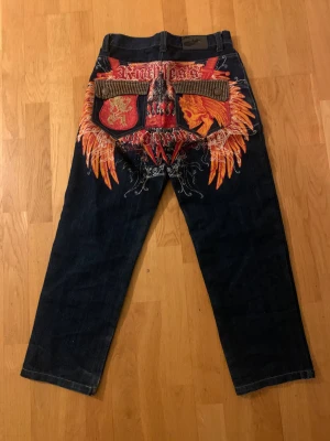 Mörkblå jeans med en snygga grafiska fickor. - Unika mörkblå jeans med rak passform och coolt broderi på bakfickorna i rött, orange och gult. Bakfickorna har detaljer med text och motiv av vingar och lejon, vilket ger ett riktigt edgy uttryck. Klassisk femficksmodell med knapp och dragkedja framtill.