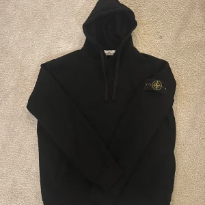 Svart hoodie från Stone Island - Svart hoodie från Stone Island med klassisk logopatch på vänster ärm. Tröjan har huva med snörning och långa ärmar. Perfekt för dig som gillar streetwear och vill ha en clean look med exklusiv känsla.