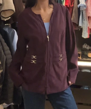 Mörklila cardigan jacka med gulddetaljer - Snygg mörklila cardigan jacka med guldfärgade dekorativa spännen vid midjan och dragkedja framtill. Jackan har lång ärm och en rak, loose passform som ger en chill vibe. Perfekt att slänga över en t-shirt för en cool look.