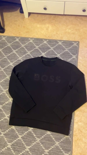 Svart tröja från BOSS med glitter - Svart tröja  från BOSS i storlek M med glittrigt BOSS-tryck på bröstet. Tröjan har rund hals, långa ärmar och en clean look med diskreta gröna detaljer på insidan av kragen. Perfekt för dig som gillar stilrena och snygga plagg.