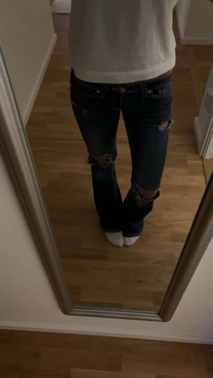 Mörkblå bootcut jeans med slitningar - Snygga mörkblå jeans med bootcut passform och slitna detaljer vid knäna och fickorna. Jeansen har vita kontrastsömmar och en broderad bakficka. Perfekt för dig som gillar en avslappnad och trendig stil.
