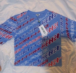 Blå Kenzo t-shirt med logomönster - Kenzo t-shirt i blått med all-over logomönster i vitt, rött och marinblått. T-shirten har rund hals och korta ärmar, tillverkad i mjuk bomull. Perfekt för dig som gillar statement-plagg och vill sticka ut med cool design.