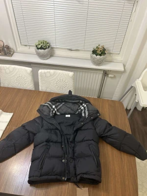 Svart pufferjacka från Burberry - Svart pufferjacka från Burberry med klassisk rutmönstrad insida vid huvan. Jackan har en snygg patch med Burberry London England på ärmen, dragkedja och knappar framtill samt två fickor med dragkedja. Perfekt för kalla dagar med sin tjocka och quiltade design.