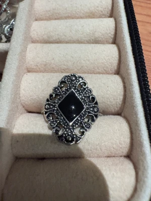 Vintageinspirerad silverring med svart sten - Snygg ring i silverfärgad metall med en stor svart, diamantformad sten i mitten. Ringen har detaljerad, ornamenterad design runt stenen som ger en cool vintagekänsla. Perfekt för dig som gillar statement-smycken med lite edge.