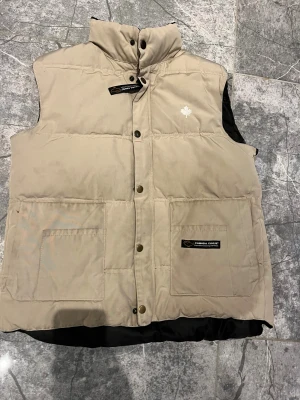 Beige dunväst från Canada Goose - Snygg beige dunväst från Canada Goose med hög krage, stora framfickor och tryckknappar. Västen har svart innerfoder och en broderad vit lönnlöv-detalj på bröstet. Perfekt för lager-på-lager och streetstyle.