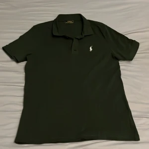Mörkgrön piké från Polo Ralph Lauren - Snygg mörkgrön pikétröja från Polo Ralph Lauren med klassisk krage och korta ärmar. Tröjan har den ikoniska vita broderade loggan på bröstet och är gjord i mjuk bomull. Perfekt för dig som gillar stilrena och tidlösa plagg.
