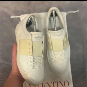 Valentino Garavani Open sneakers vit - Snygga Valentino Garavani Open sneakers i vitt skinn med en ljusbeige panel över mitten och ikoniska nitar runt hälen. Klassisk rund tå, platt sula och vita snören. Perfekta för dig som vill ha en clean och lyxig look med en twist. Det är lite märke på skon men inget som syns när man bär dem, dust bag följer med.