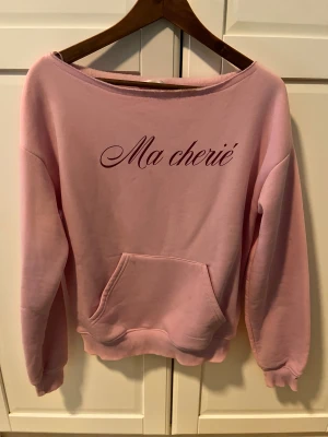 Rosa sweatshirt med text Ma cherié - Säljer en ljusrosa sweatshirt med snyggt tryck 'Ma cherié' framtill. Tröjan har en bred halsringning, långärmade ärmar och en stor magficka. Perfekt för dig som gillar chill och trendiga plagg med fransk vibe.