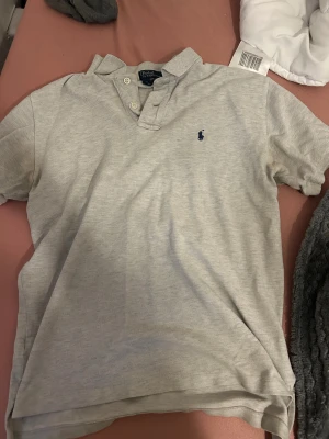 Ljusgrå pikétröja från Ralph Lauren - Snygg ljusgrå pikétröja från Polo Ralph Lauren i storlek M. Tröjan har klassisk krage, korta ärmar och två vita knappar vid halsen. Ikoniska blå Polo-loggan broderad på bröstet. Tillverkad i mjuk bomull som är skön mot huden. Storlek M för 12-14 åringar. tröjan har ett litet litet hål men det är knappt synbart när man har på sig tröjan. så klart så går priset att diskutera🤩