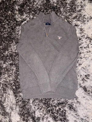 Grå stickad half zip-tröja GANT - Snygg grå stickad tröja från GANT med half zip och hög krage. Tröjan har långärmade ribbade muddar och broderad logga på bröstet. Perfekt för dig som gillar klassisk stil med en modern touch.