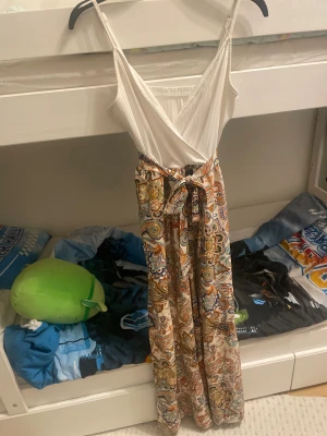 Paisleymönstrad byxdress med omlott - Säljer en snygg byxdress med paisleymönstrade vida byxben i beige, orange, blå och gröna toner. Toppen är vit, ribbad och har omlott med smala axelband. Midjan har ett knytband för extra stilpoäng. Perfekt för dig som gillar boho-vibe och färg!