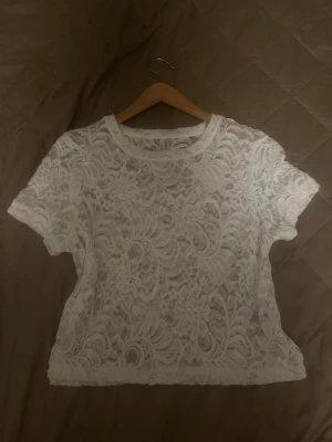 Vit spetsblus med kort ärm - Säljer en vit blus från Monki i genombruten spets med blommigt mönster. Modellen är croppad med rund halsringning och korta ärmar. Perfekt för dig som gillar romantiska detaljer och vill ha en luftig topp med snygg struktur.